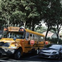 As students exit a school bus, which has its red "Stop Sign" extended, blurred vehicles illegally speed past the bus instead of stopping, putting the students in danger.
