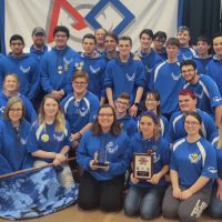 High School Robotics Team dressed in blue shirts holding their awards, a plaque and a crystal award.